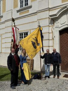 Fahnenweihe der Metalltechnik-Innung 2 Vor dem historischen Gebäude mit seiner barocken Fassade präsentieren vier Teilnehmer stolz zwei traditionsreiche Fahnen. Im Zentrum steht eine leuchtend gelbe Fahne mit einer aufwendig gestickten Darstellung von Handwerkern und der Aufschrift "Meister Egers & Lehrjünger Flums". Die Personen tragen festliche und traditionelle Kleidung und strahlen bei sonnigem Wetter eine feierliche und zugleich fröhliche Stimmung aus. Die Szene vermittelt das lebendige Bewahren von Handwerks- und Vereinsgeschichte.